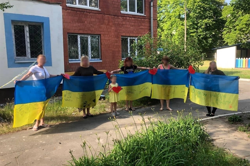 [ai] A group of six individuals holding Ukrainian flags with red and yellow hearts attached, standing in front of a building surrounded by greenery. The scene depicts community support and patriotism.